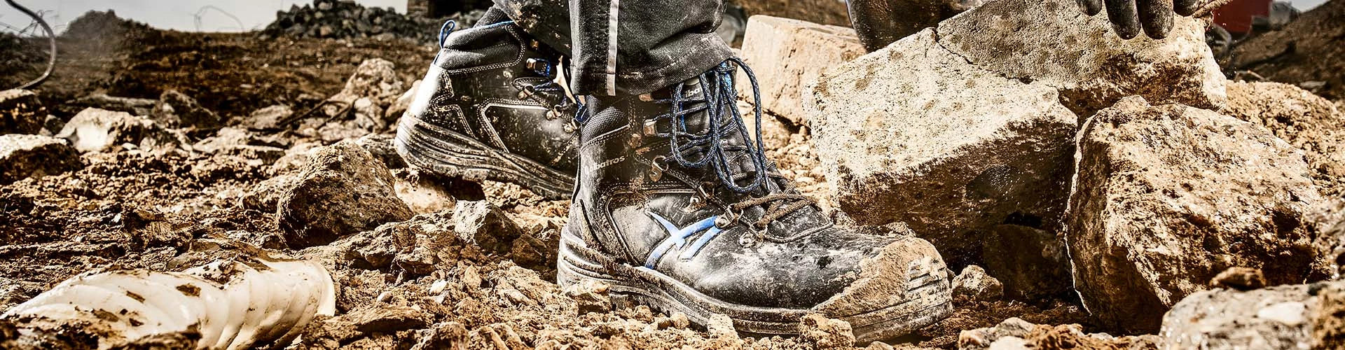 Robuste ALBATROS UNIT BAU MID Sicherheitsschuhe S3 in Schwarz mit blauen Details, stark verschmutzt im Einsatz auf einer schlammigen Baustelle zwischen Steinen und Geröll