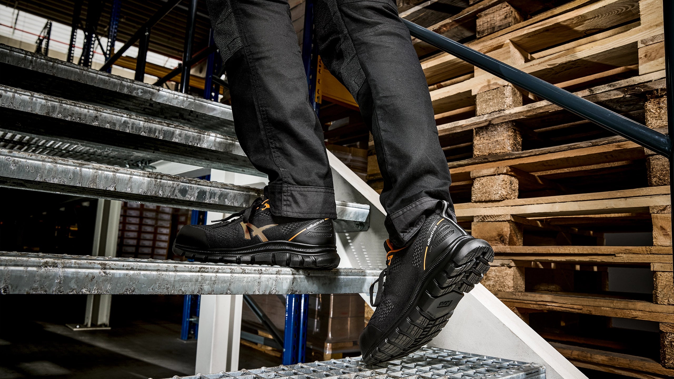 Ein Paar schwarze Albatros LEVITATE ORANGE LOW S1PS SR Sicherheitsschuhe wird auf einer Metalltreppe in einem Lagerumfeld getragen.