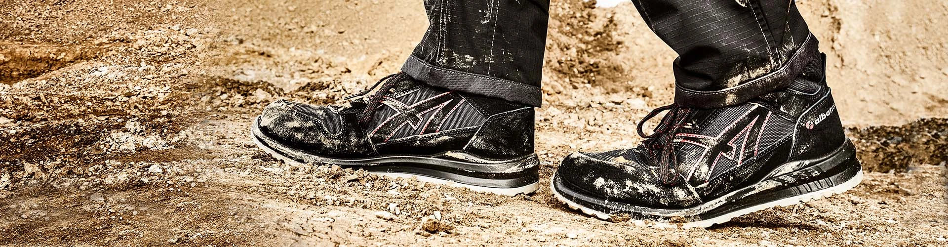 Ein Paar schwarze Albatros-Sicherheitsschuhe mit roten Akzenten wird auf einer staubigen, unebenen Erdoberfläche getragen.