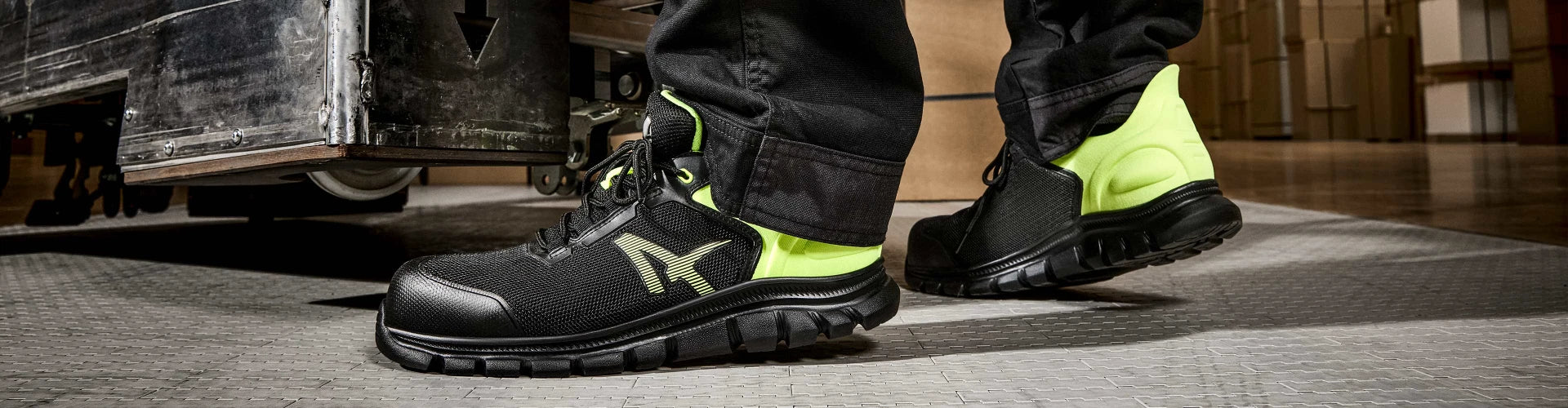 Schwarze ALBATROS OCTANE YELLOW LOW Sicherheitsschuhe S1PS mit gelben Akzenten, getragen auf einer Industriehalle-Bodenplatte neben einem Hubwagen