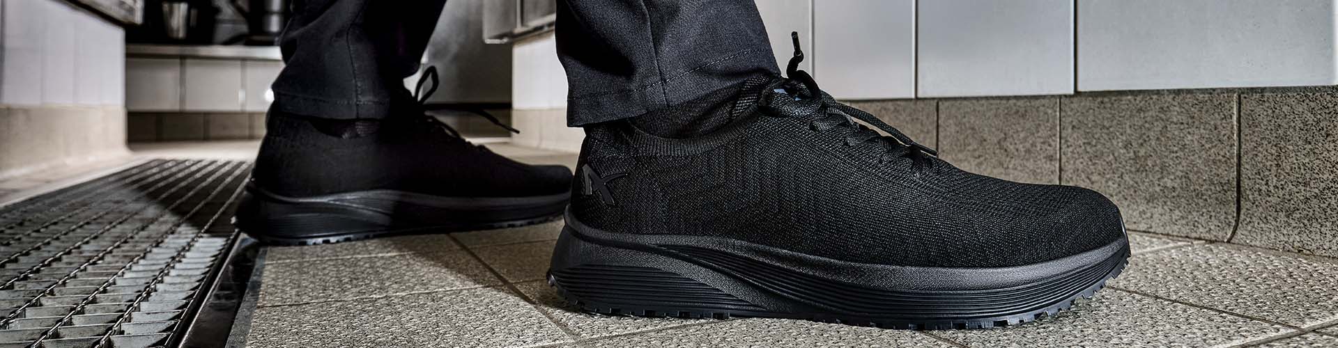 Ein Paar schwarze Albatros MOOD ST BLK LOW Schuhe wird in einer gewerblichen Küche auf hellen Fliesen getragen.