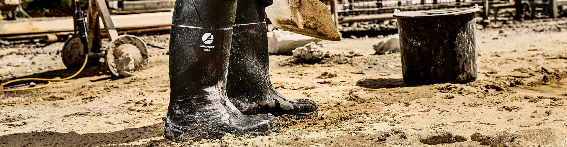 Ein Paar schwarze Albatros TITAN S5 FO CI SR PU-Sicherheitsstiefel steht im sandigen Baustellenbereich, sichtbar verschmutzt vom Arbeiten.